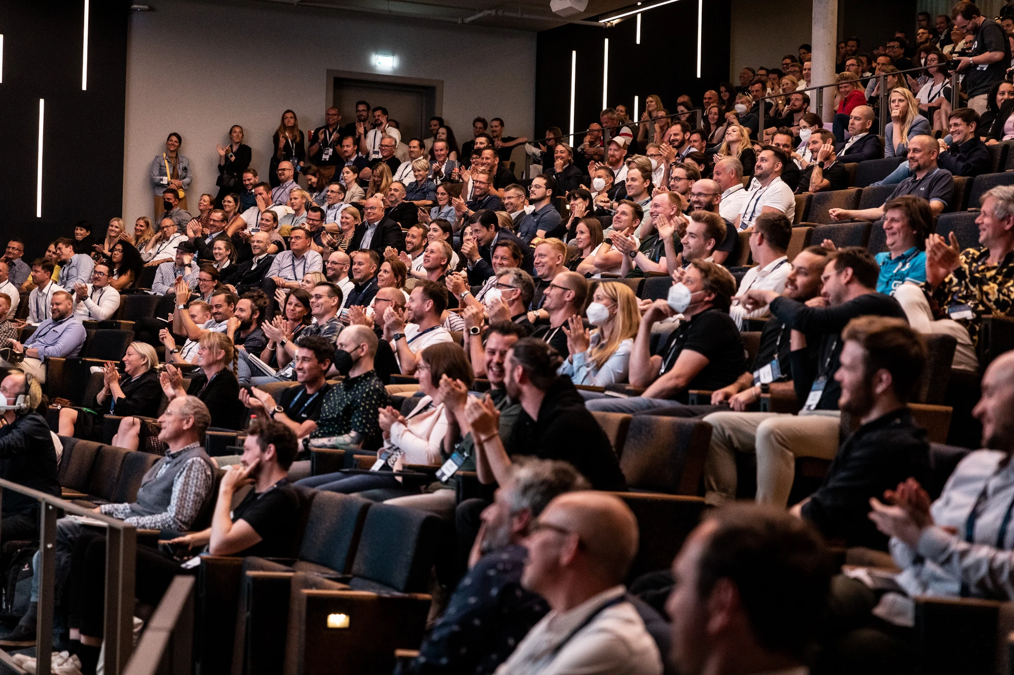 Ein voll besetzter Publikumsbereich auf der K5. Zahlreiche Teilnehmerinnen und Teilnehmer sitzen in dunklen Polstersesseln eines auditoriumsähnlichen Saals. Sie blicken gebannt nach vorne zur Bühne, lächeln und applaudieren begeistert. Das Bild fängt die positive, inspirierende Atmosphäre der K5 Konferenz ein.