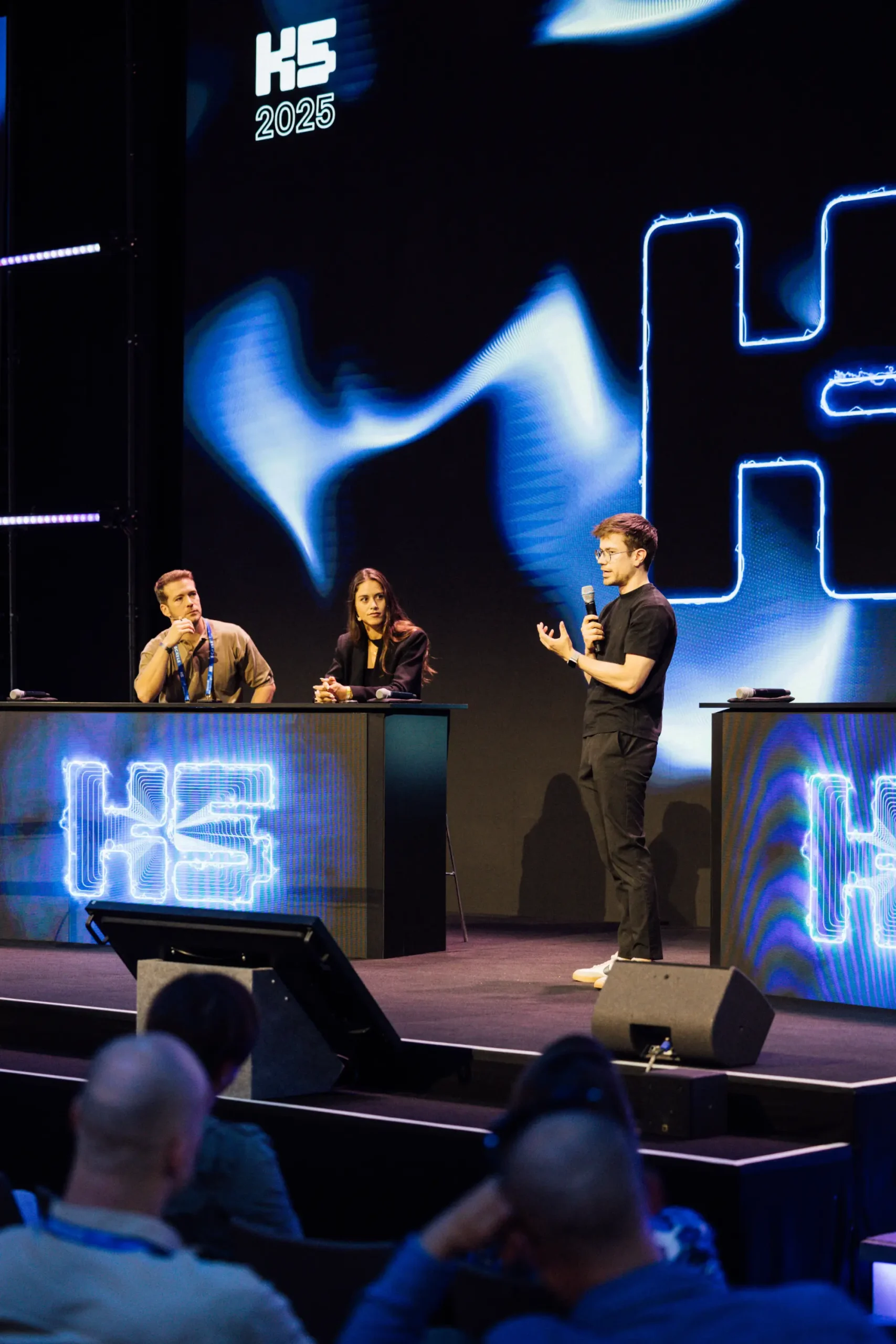 Ein Gründer steht im schwarzen Outfit auf der großen, dunkel gehaltenen Bühne der K5 Konferenz und spricht konzentriert in ein Mikrofon. Links von ihm sitzt eine zweiköpfige Experten-Jury an einem Rednerpult, das mit leuchtend blauen K5-Logos gebrandet ist, und hört dem Pitch aufmerksam zu. Im Vordergrund sind unscharf die Köpfe des Publikums zu erkennen.